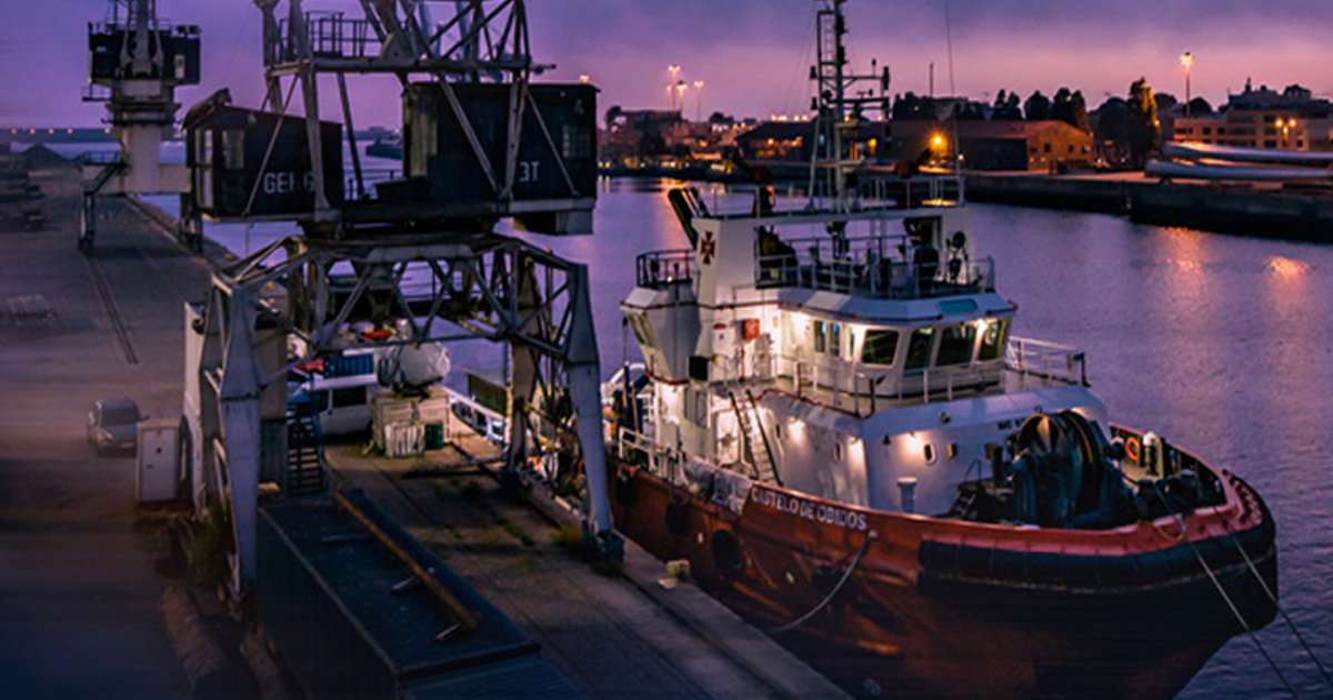 Tugboat docked under a harbor crane at dusk.