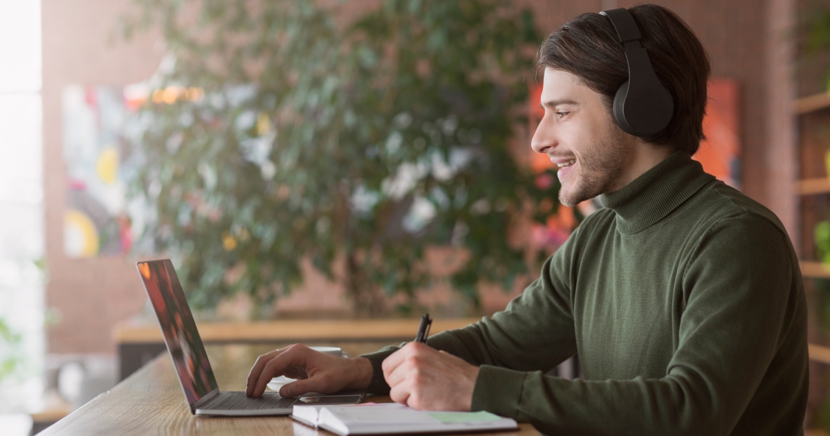 Smiling man with headphones taking notes at a laptop.