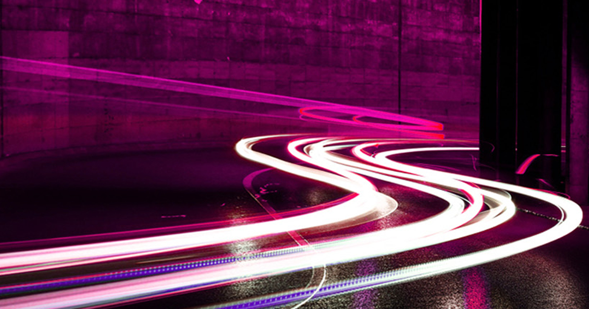 Long-exposure light trails sweeping through a dark curve in pink and white, representing high-speed performance engineering.