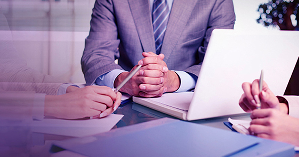 Insurance advisor with folded hands meeting two clients across a desk with a laptop, representing the insurance industry during COVID-19.