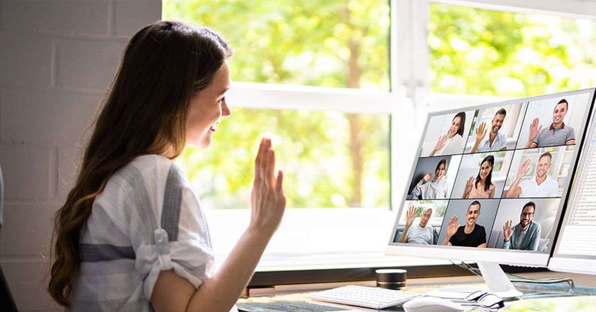 Student waving to classmates in a video-call grid on her monitor during a remote lesson, representing remote learning during COVID-19.