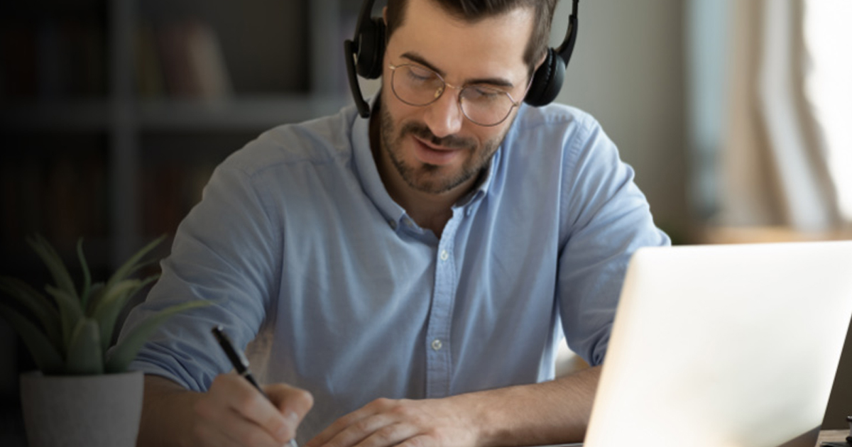 Man wearing a headset taking notes in front of a laptop during an online course, representing upskilling and reskilling remotely.